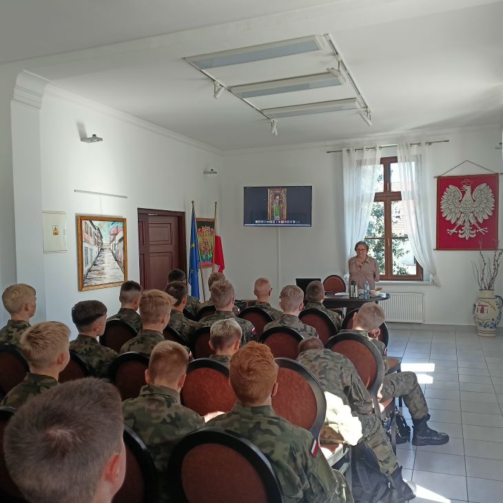 Grupa kilkunastu młodych osób w mundurach siedzi na krzesłach w pomieszczeniu. Dorosła kobieta stoi do nich przodem. Na ścianie widoczne godło Polski.